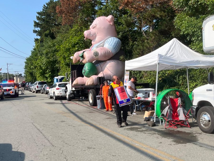 A large inflatable greedy pig with a green money bag sits on a flatbed truck, next to those involved in the Teamsters Local 251 Union strike at DHL Express ServicePoint in Pawtucket.